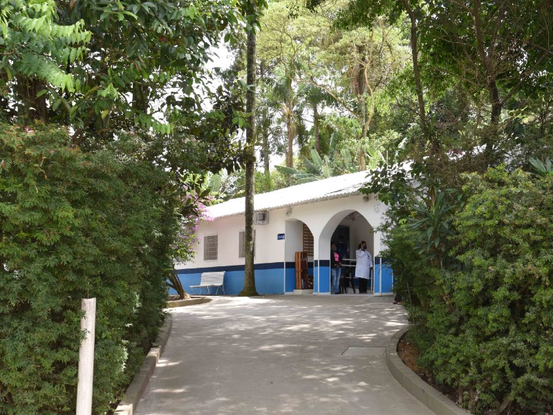 Entrada da UBS Chácara Santa Maria, com paredes brancas e azuis, ao fundo, entre um caminho com plantas dos dois lados. Duas pessoas estão paradas na entrada